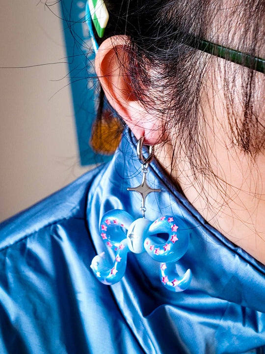 Close-up of a person wearing an ice-blue snake bowtie earring with pink stars and a star-shaped charm.